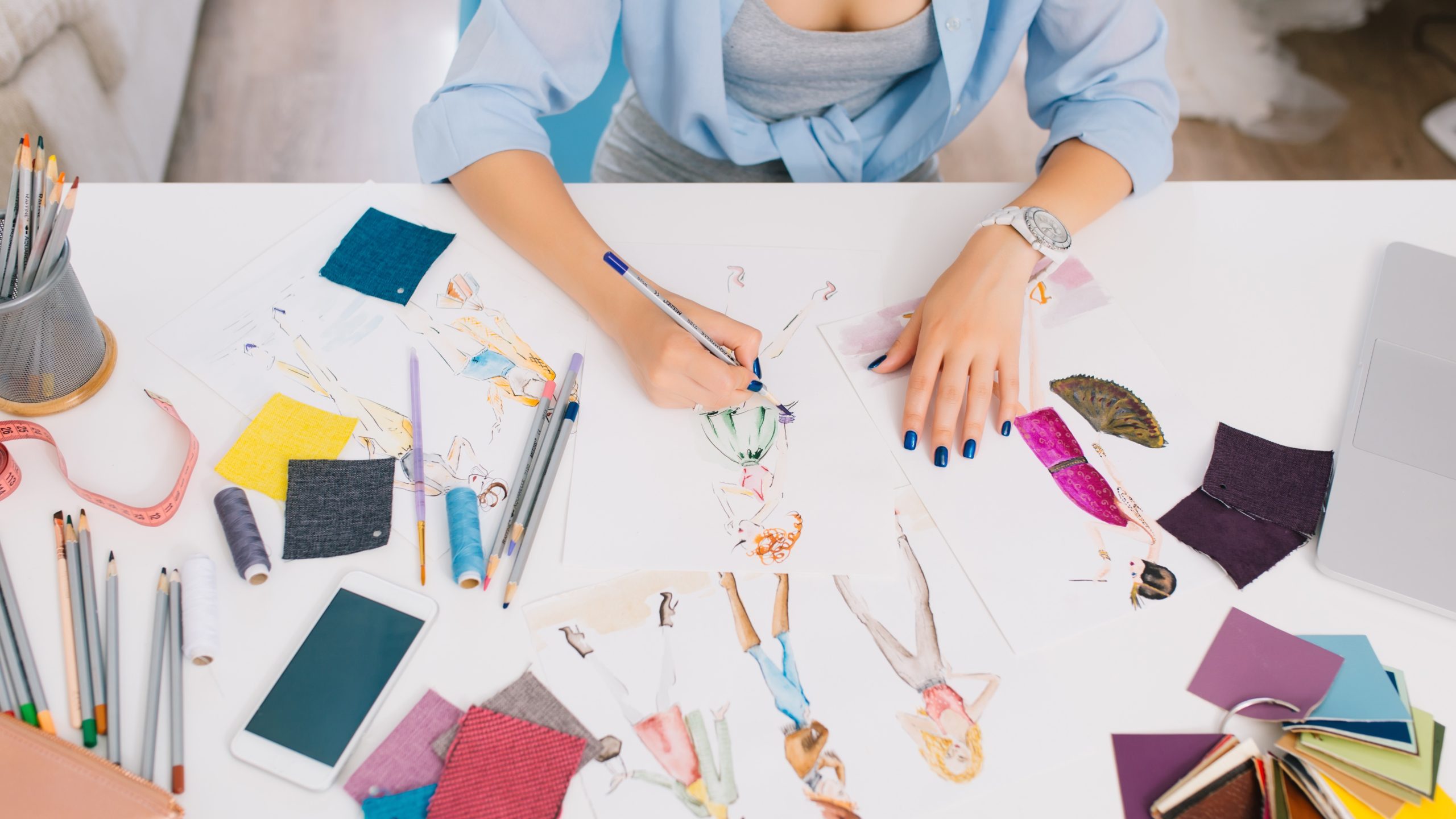 This picture describes the processes of designing clothes. There are hands of a girl drawing sketches on the table. There is creative mess with different stuff on the table.